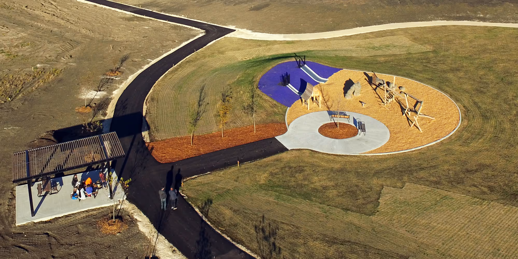 Aerial photograph of a nature playground on a hill with a circular sand and wood chip play area, a blue slide mound, a shade pergola and a winding path.