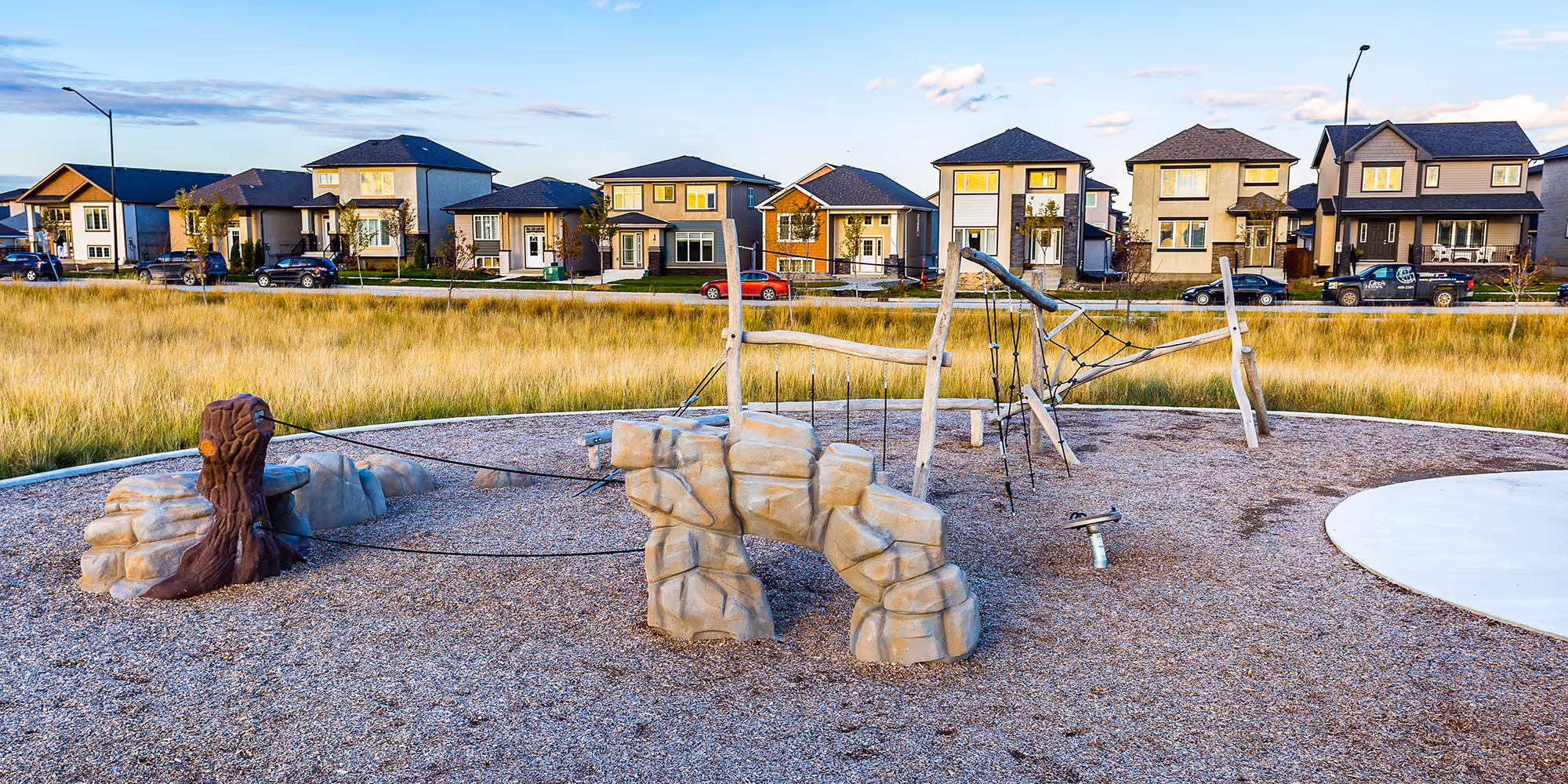 Playground with stone-textured climbing forms, a rope net frame and a tree stump on wood chips, with a row of houses and tall grass behind.