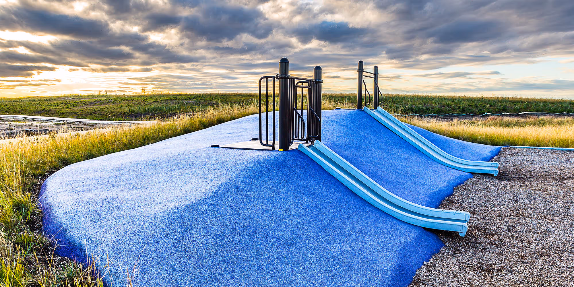 Blue rubber surfaced slide mound with two blue slides and a black climbing frame, set in tall grass under a cloudy sunset sky.
