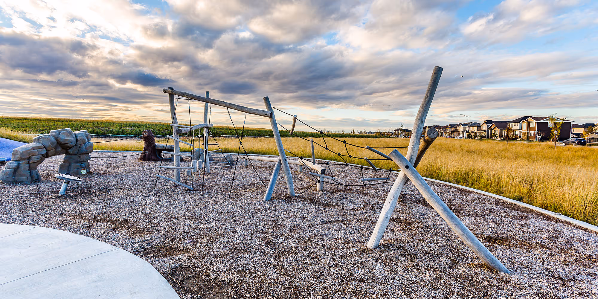 Timber log rope climbing frame and a stone arch climbing feature on wood chips, with a curved concrete edge, tall grass and houses on the horizon.