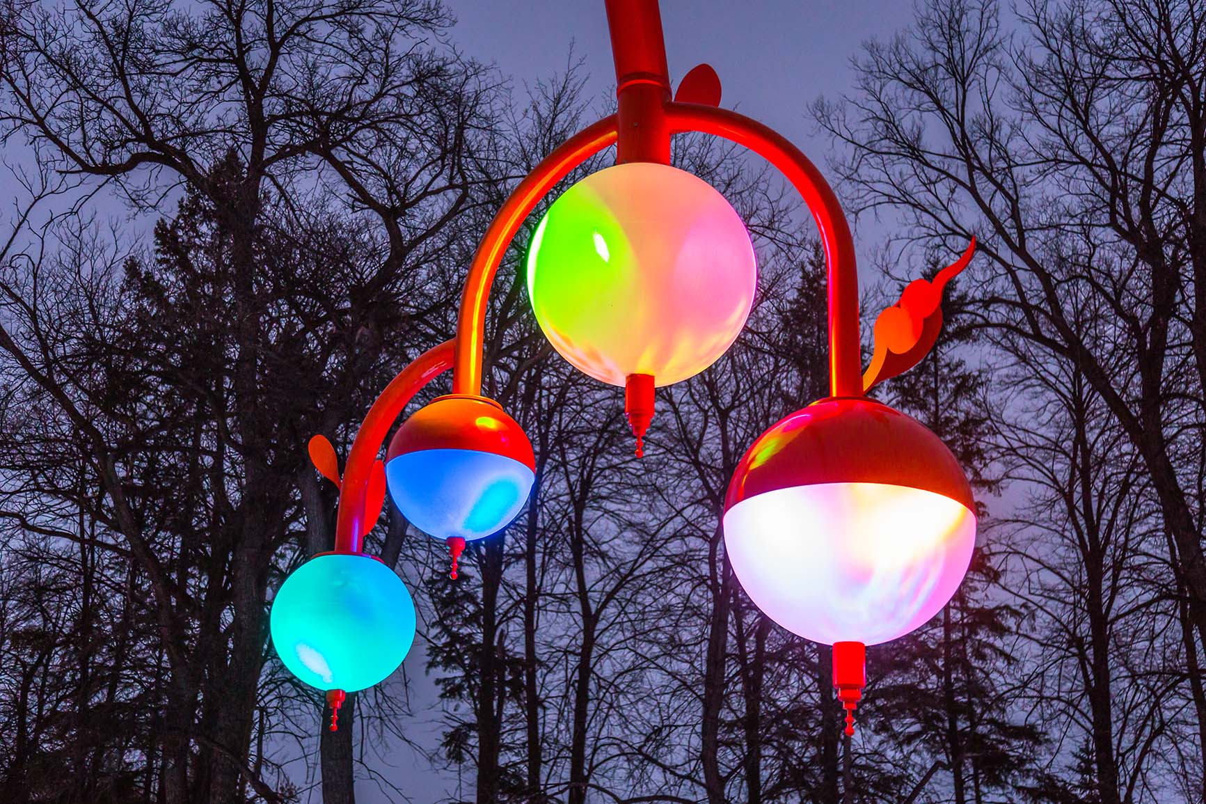 Close-up at dusk of an orange curved pole holding four glowing spherical lights in green, blue, pink and white, with bare trees behind.
